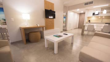 A modern living room with a white coffee table, a TV mounted on a wooden panel, and a kitchen area in the background.