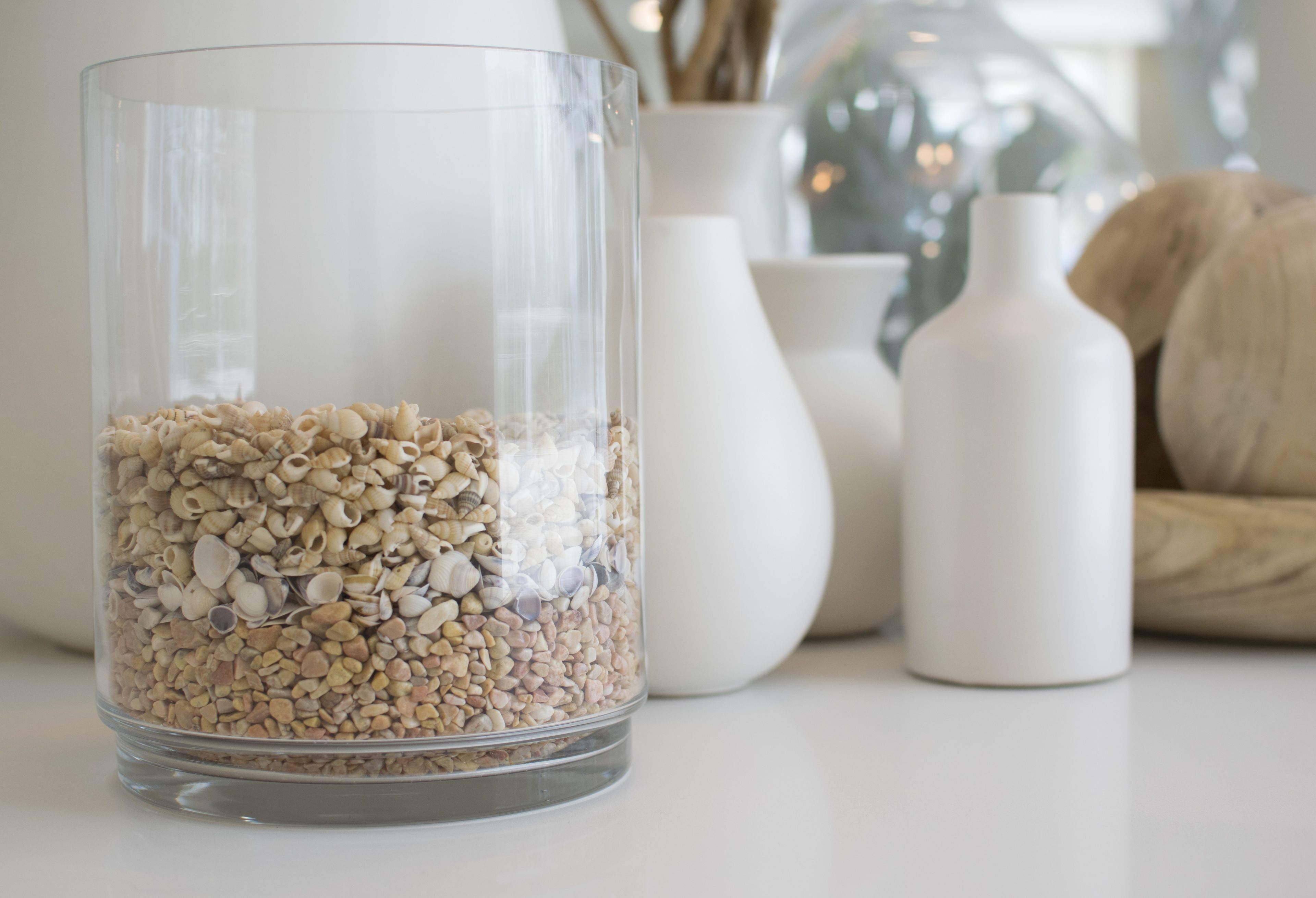 Een transparante glazen vaas gevuld met diverse schelpen, geplaatst op een witte ondergrond met witte vazen en een houten schaal op de achtergrond.