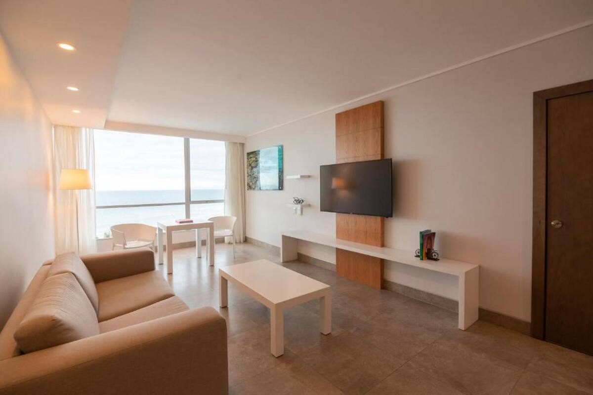 Una sala de estar moderna con un sofá, una mesa de centro, un televisor instalado en la pared y un ventanal con vista al mar.