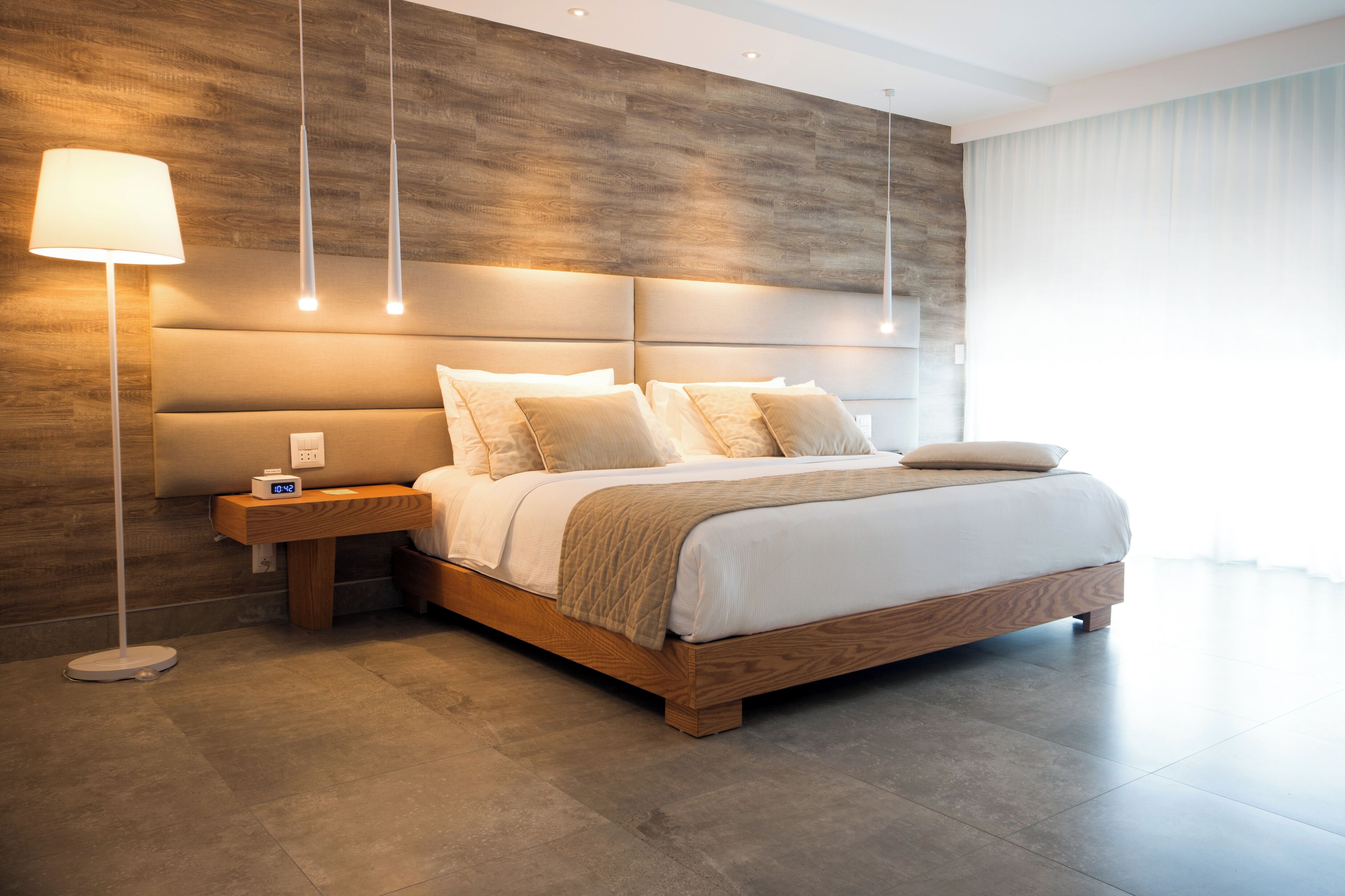 A modern bedroom with a wooden headboard, a bedside table with a lamp, and a large bed with multiple pillows.
