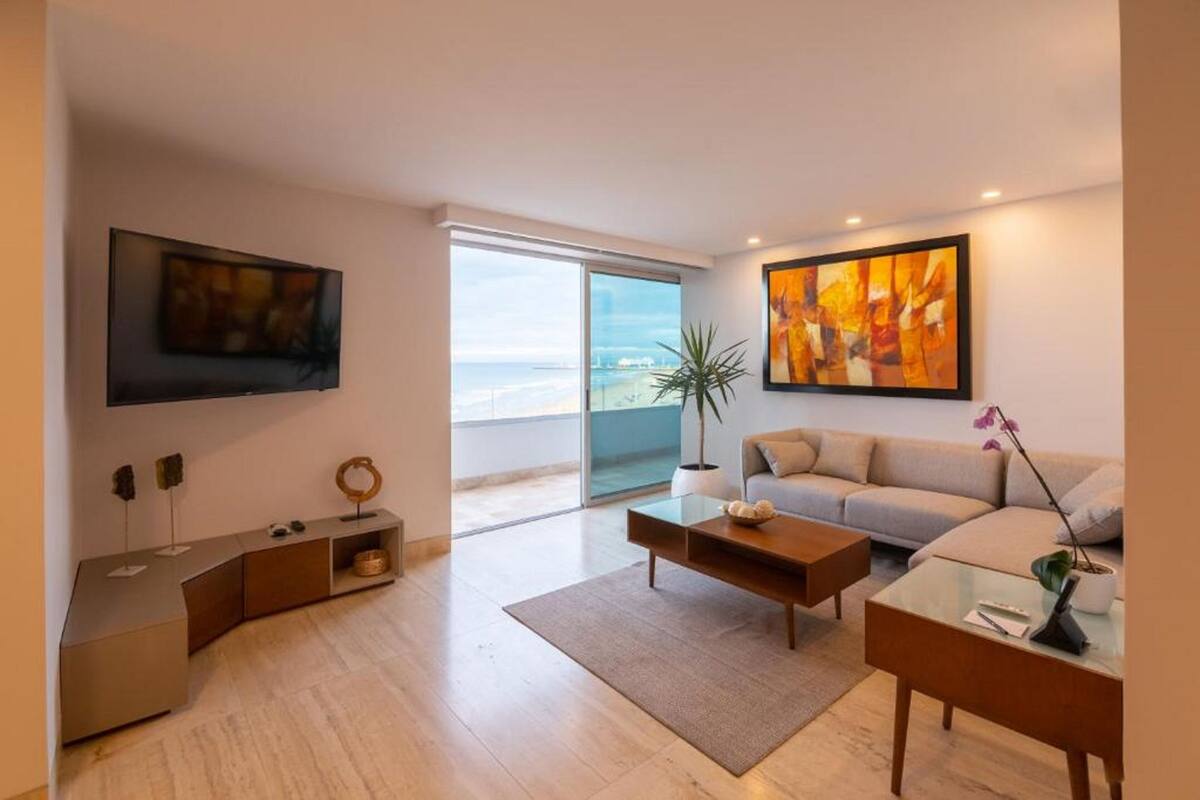 Una sala de estar moderna con un sofá, una mesa de centro y un televisor. Hay un ventanal con vistas a la playa.