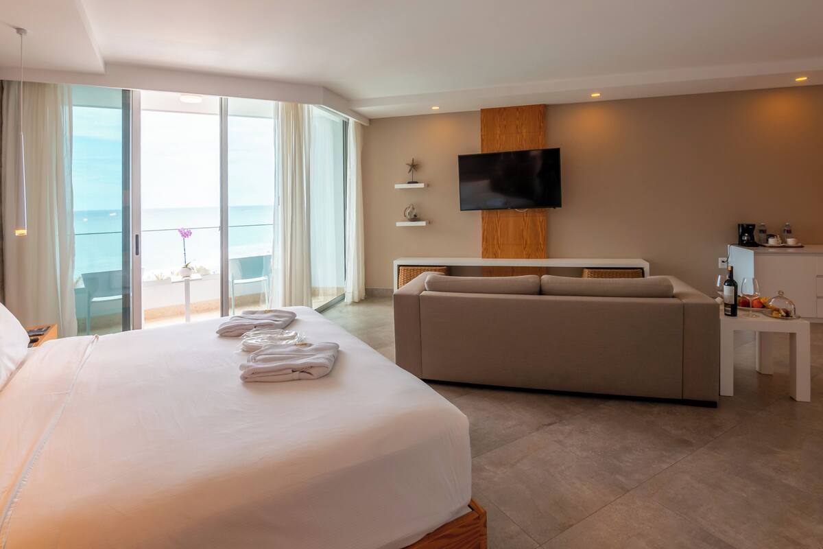Habitación de hotel con una cama grande, un sofá, un televisor, un balcón con vistas y una kitchenette.