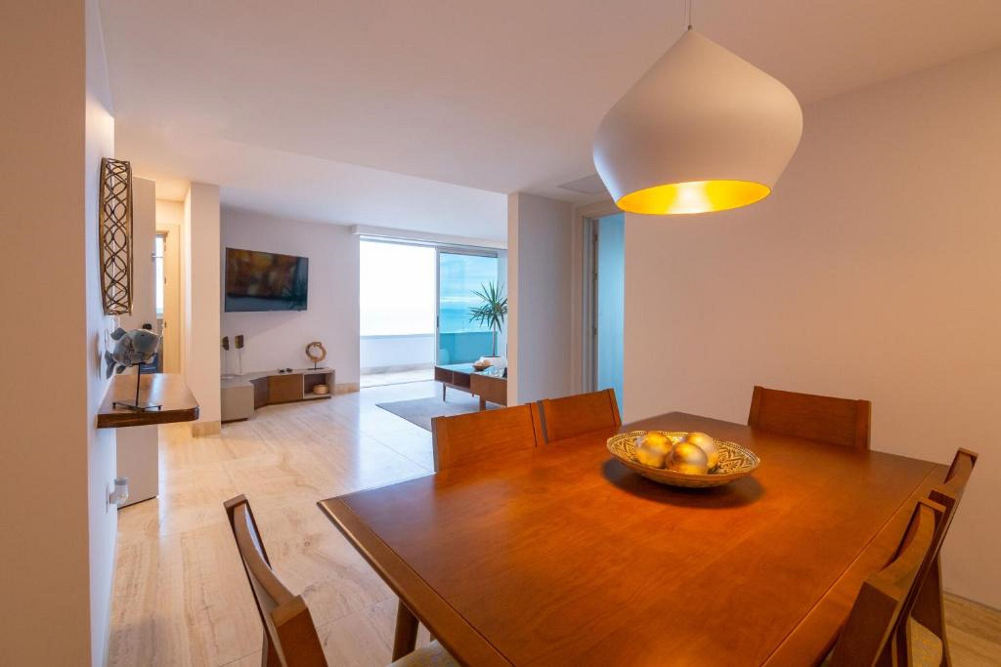 A modern dining area with a wooden table, chairs, and a bowl of fruit. There is a pendant light above the table.