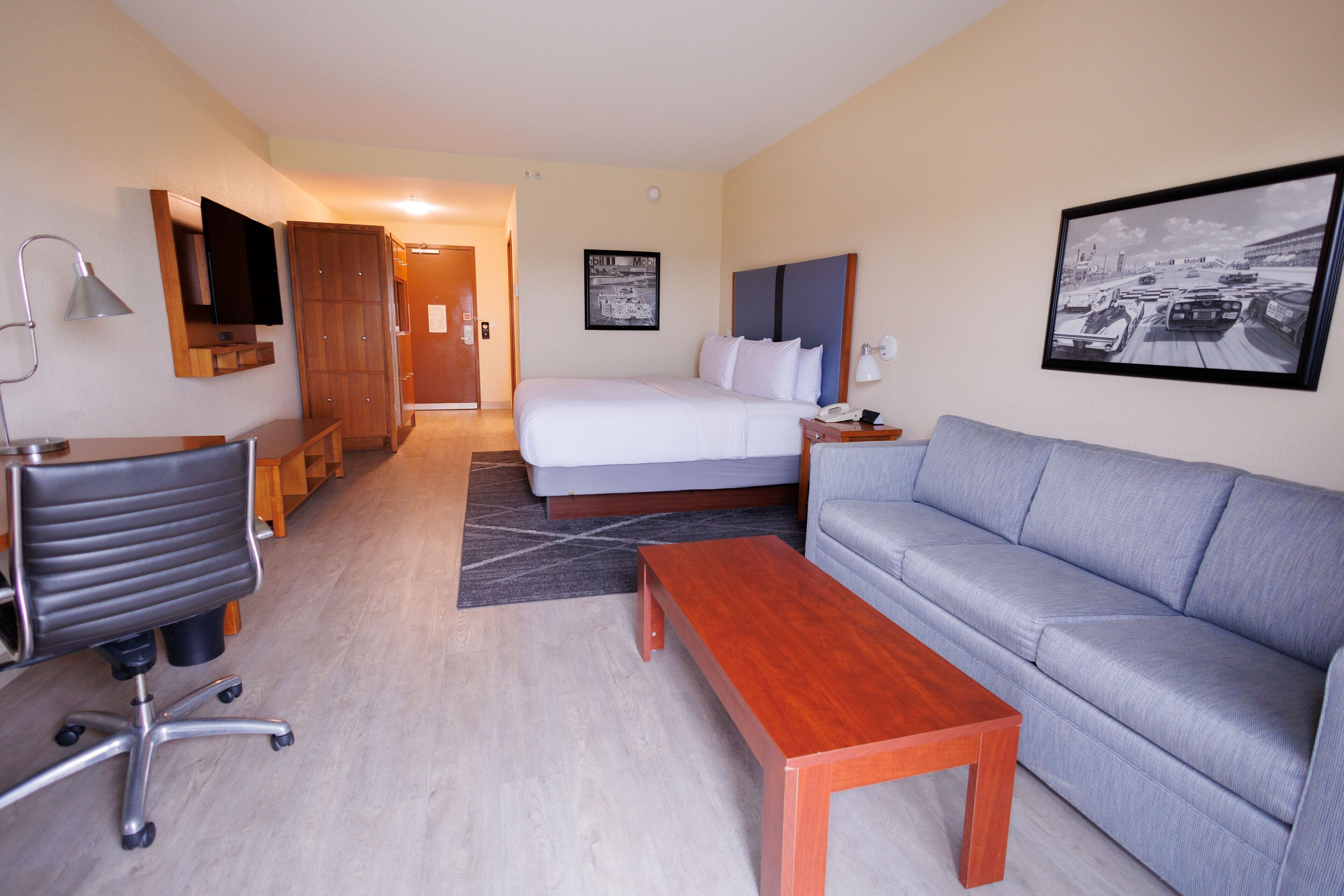 A hotel room with a bed, a sofa, a desk with a chair, a wooden coffee table, a TV mounted on the wall, and a framed picture on the wall.