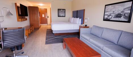 A hotel room with a bed, a sofa, a desk with a chair, a wooden coffee table, a TV mounted on the wall, and a framed picture on the wall.