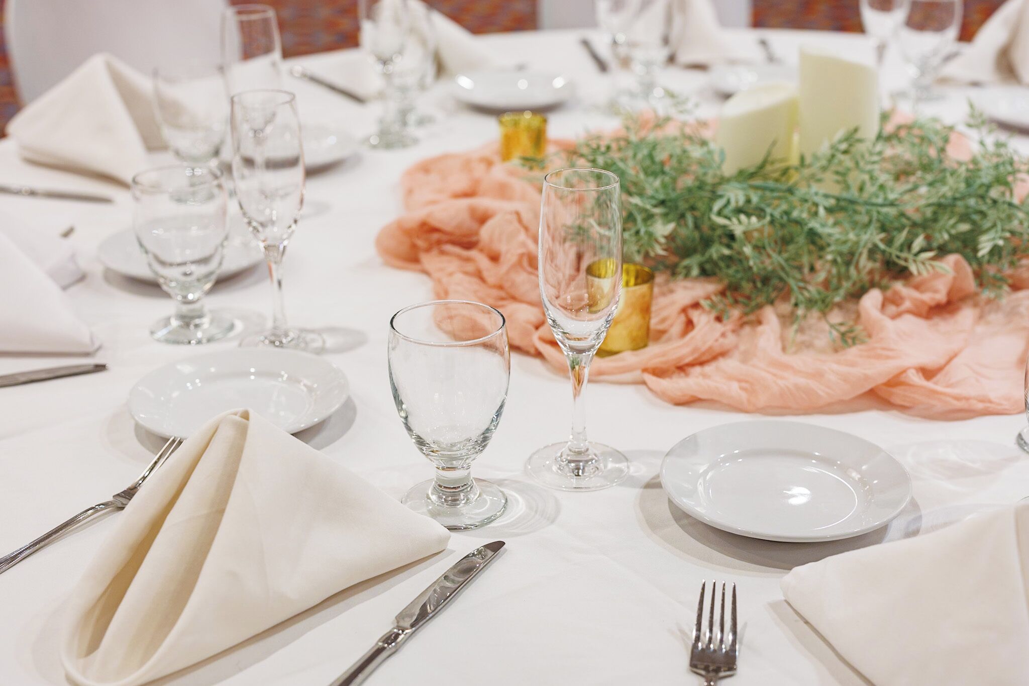 Une table dressée avec de l’argenterie, de la vaisselle blanche et des verres transparents, agrémentée d’un tissu rose et de quelques éléments de verdure.