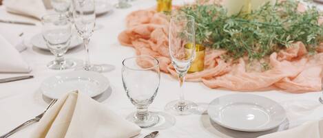 Une table dressée avec de l’argenterie, de la vaisselle blanche et des verres transparents, agrémentée d’un tissu rose et de quelques éléments de verdure.