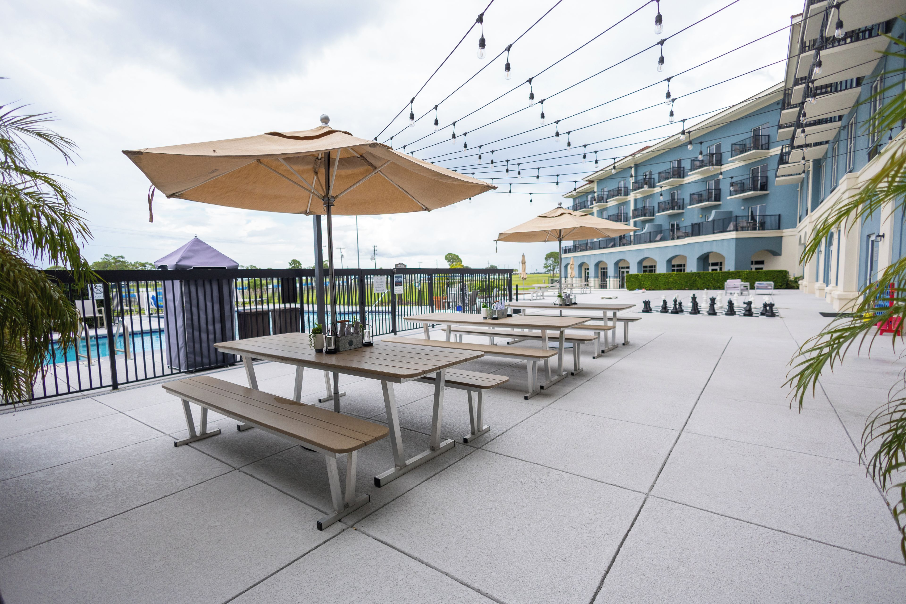 An outdoor patio area with tables and benches, umbrellas, and a pool in the background.