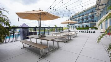 Un patio al aire libre con mesas, bancos, sombrillas y una piscina al fondo.