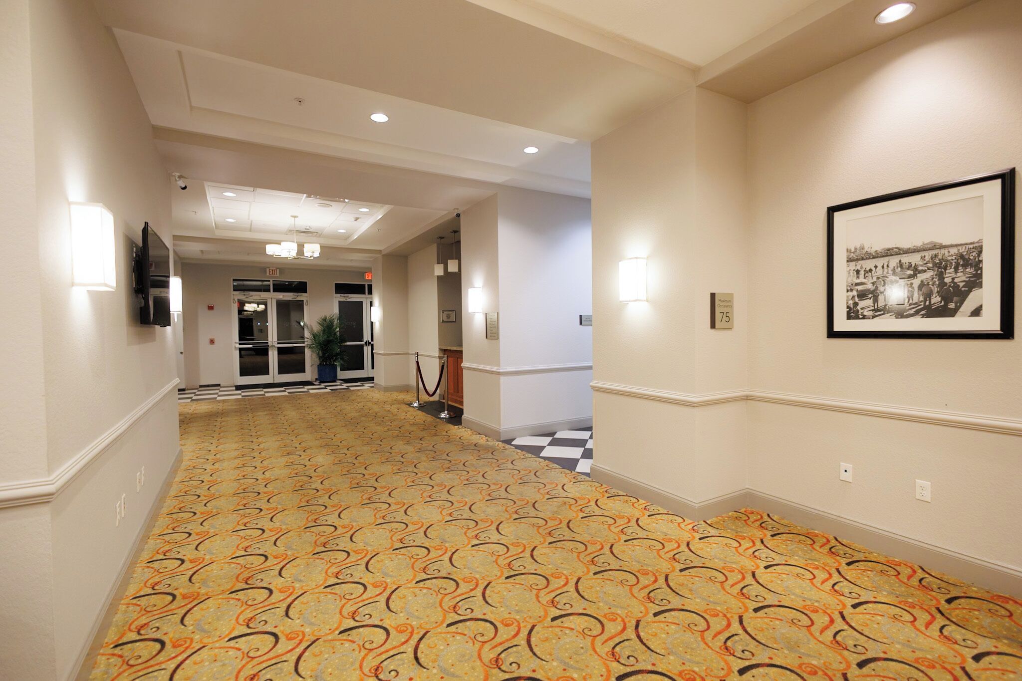A hotel corridor with patterned carpet, recessed lighting, a framed photograph on the wall, and a checkered floor pattern.