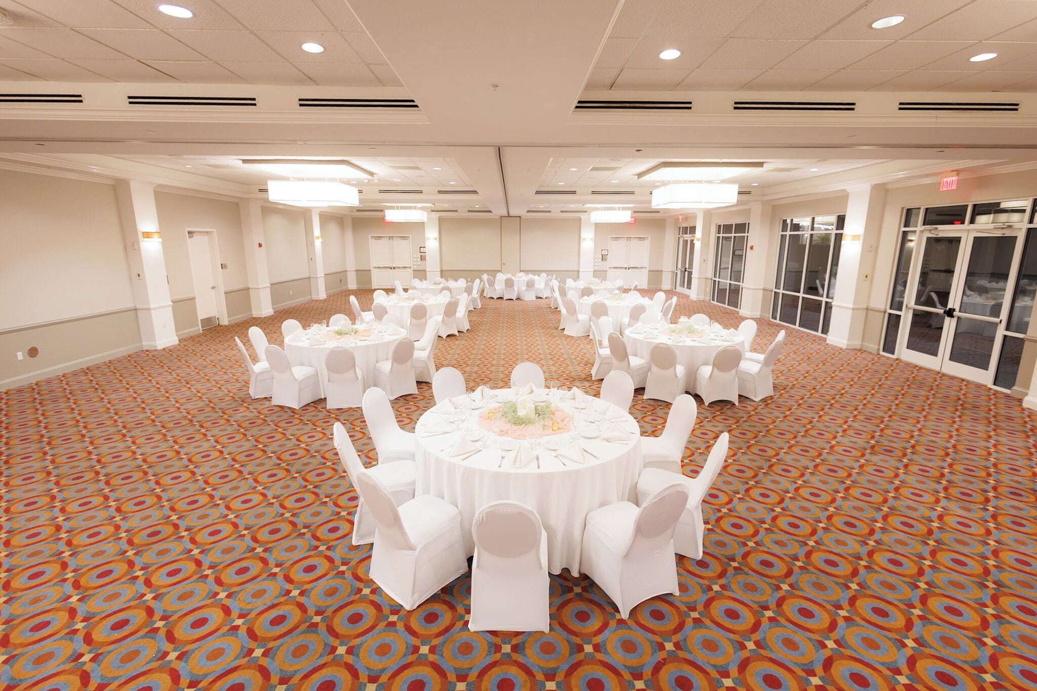 A large, circular banquet hall with white round tables and chairs, carpeted floor, and large windows.