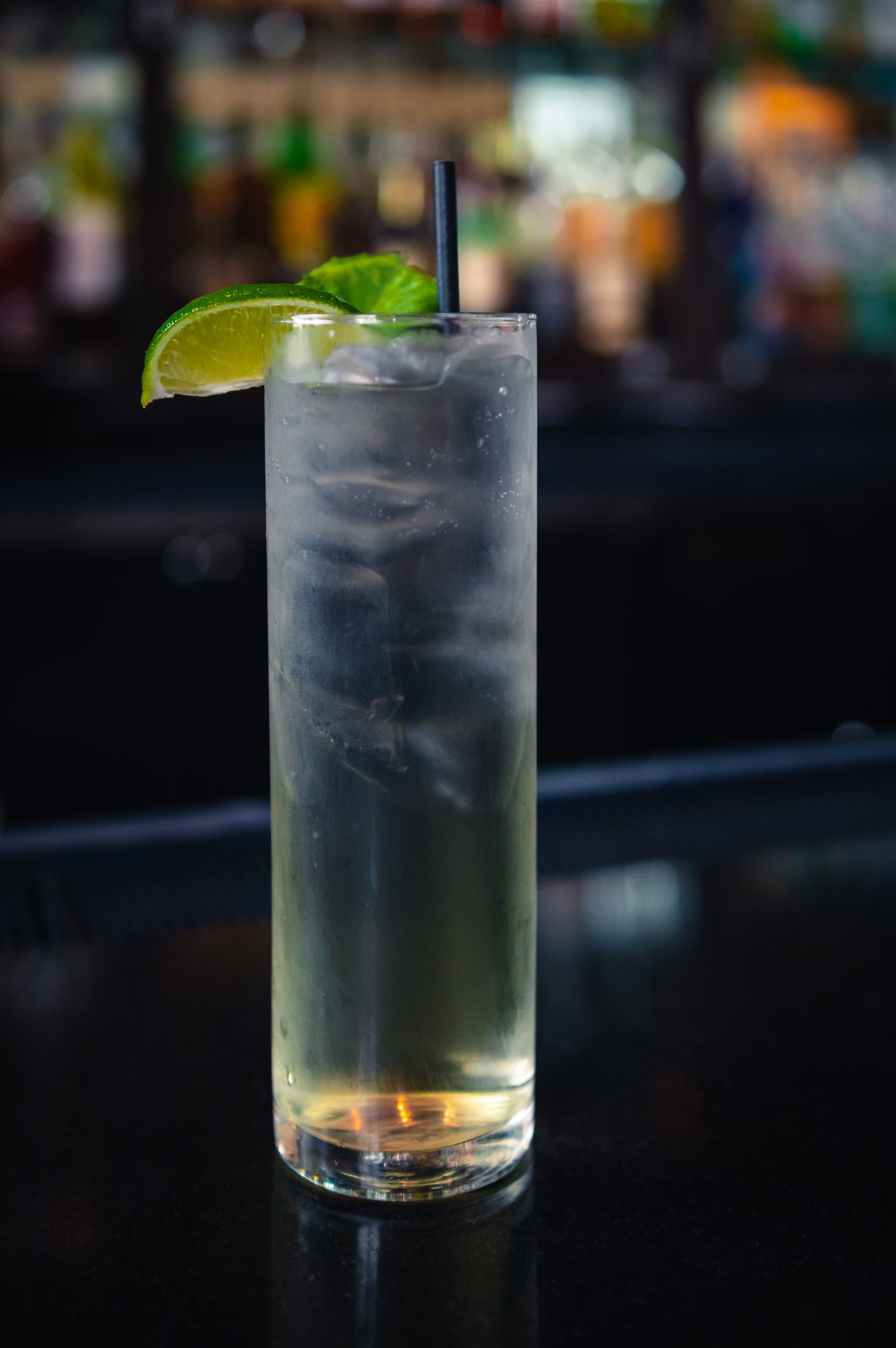 A tall glass with a lime wedge and a straw, filled with a clear liquid and ice cubes, on a bar counter.