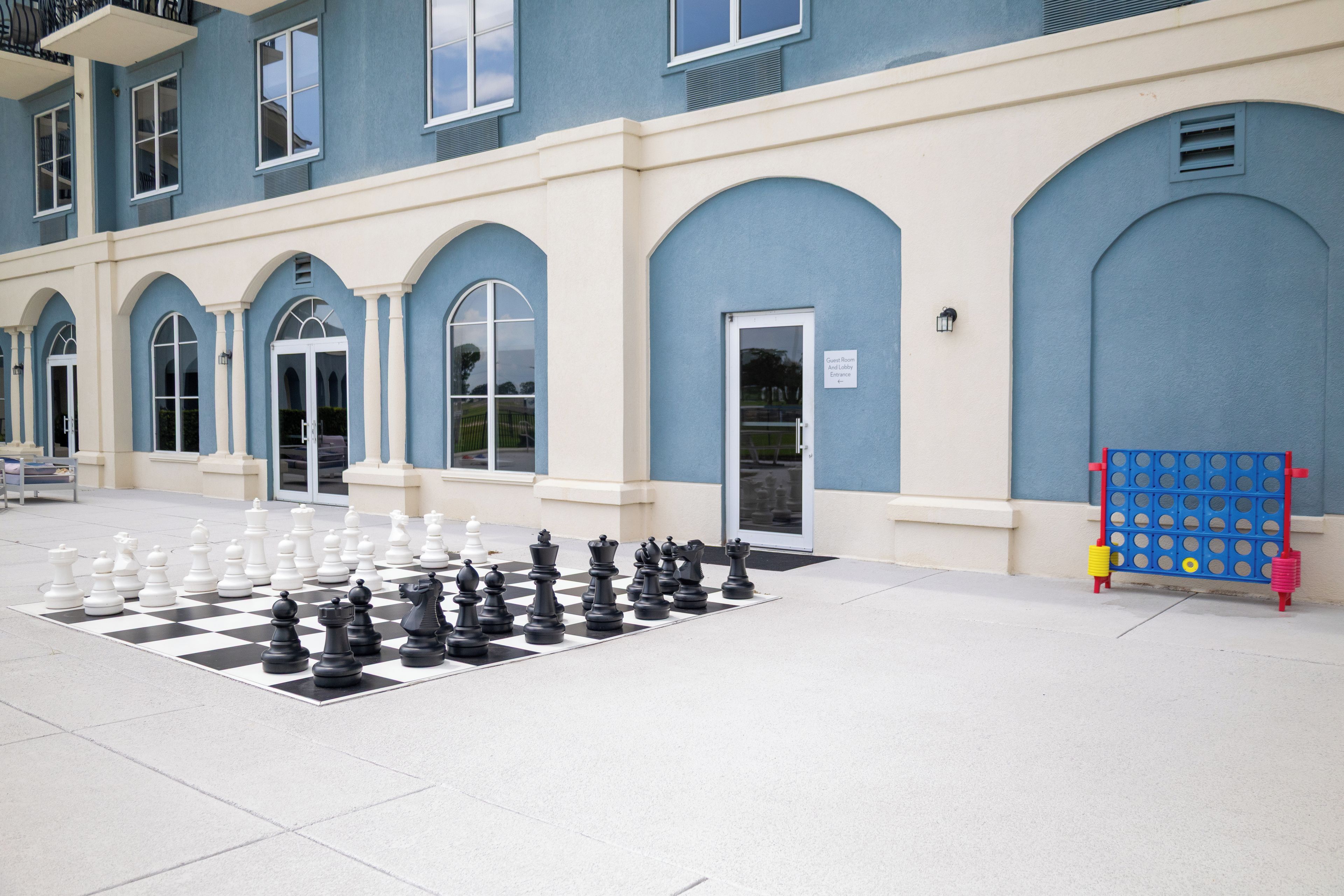 Un jeu d’échecs est installé sur une surface pavée devant un bâtiment doté de fenêtres en arc de cercle et d’une enseigne indiquant l’entrée des chambres et du hall d’accueil.