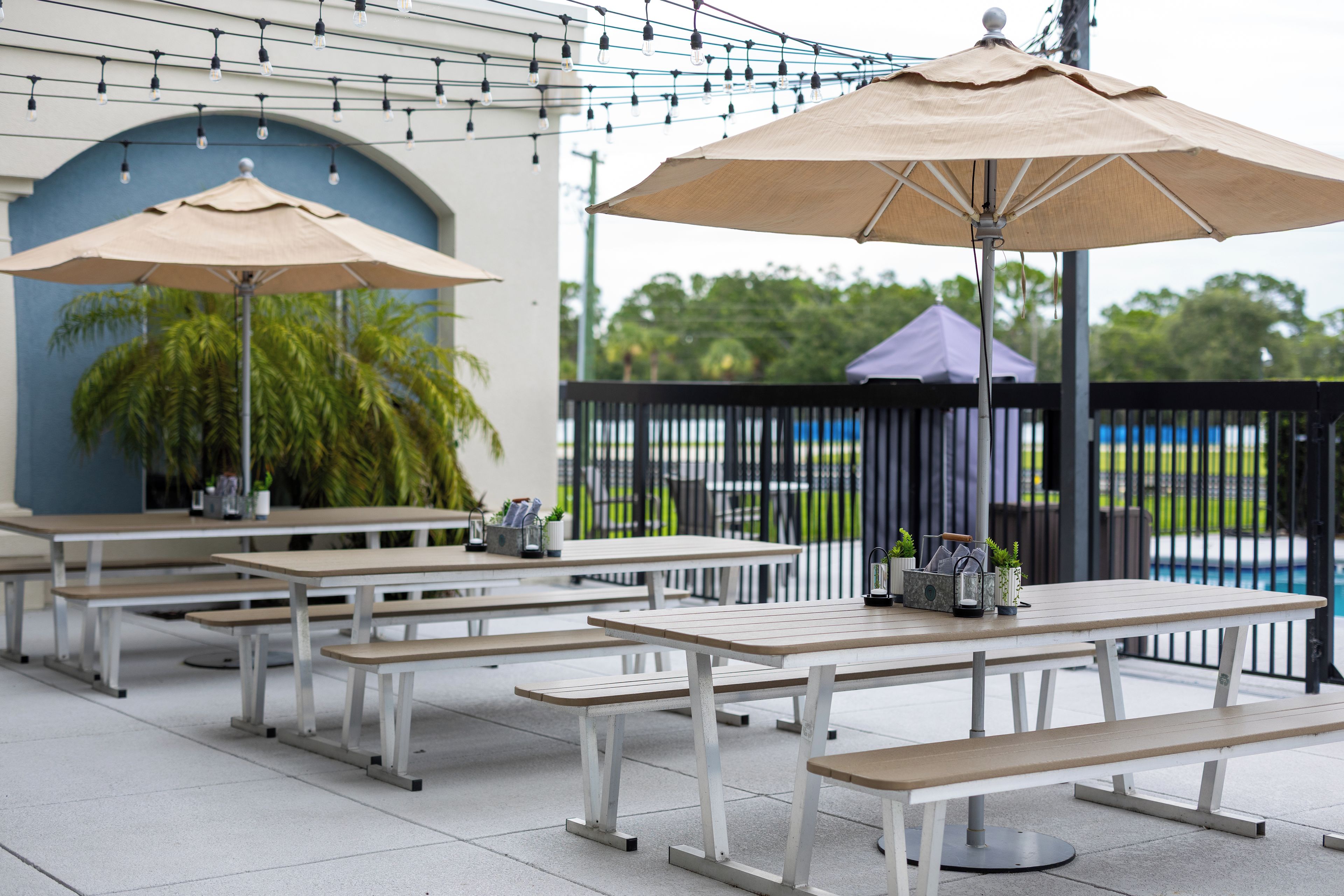 Outdoor seating with umbrellas, tables, and benches, with a pool and greenery in the background.