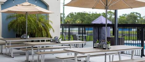 Outdoor seating with umbrellas, tables, and benches, with a pool and greenery in the background.
