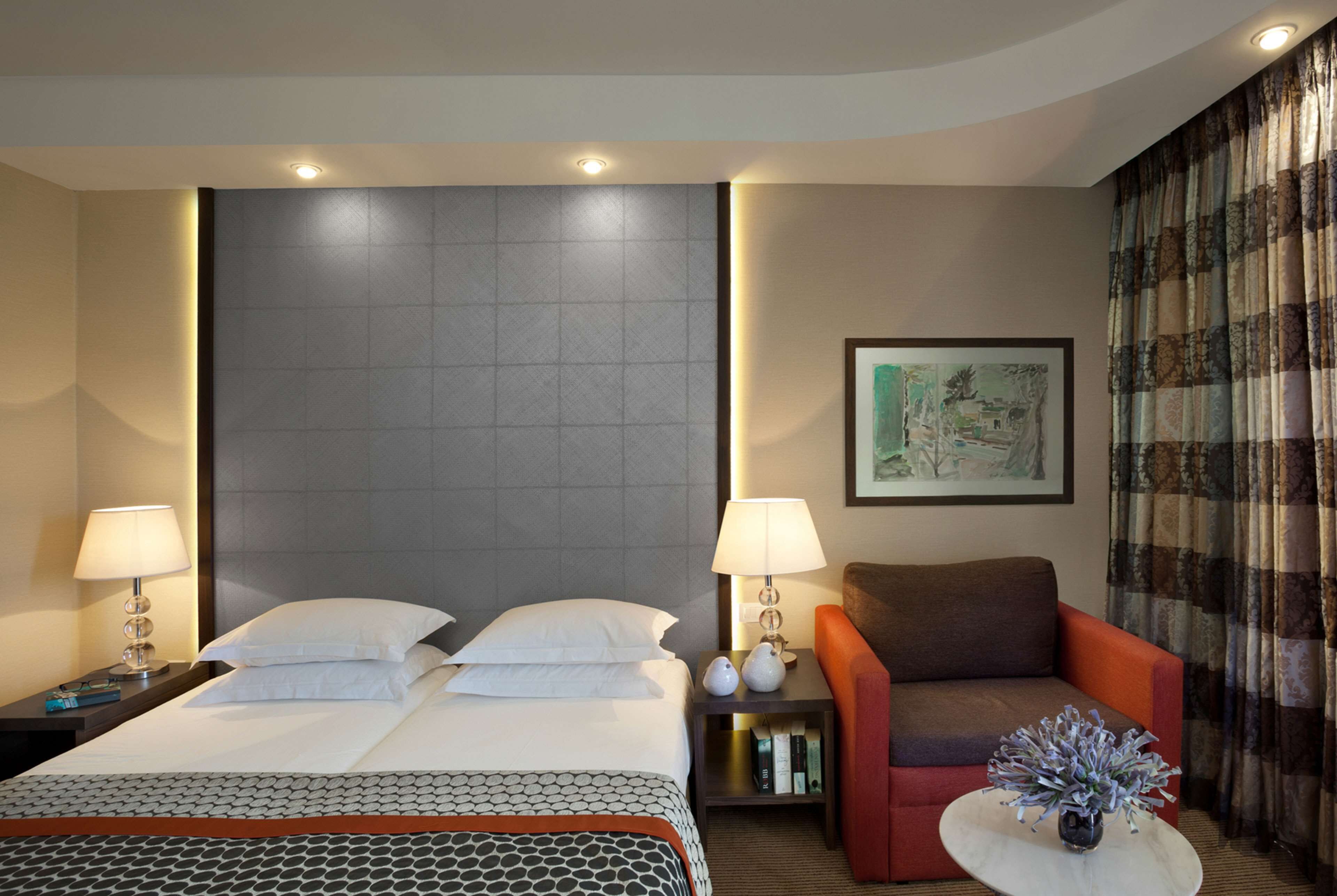 Une chambre d’hôtel avec un grand lit, deux tables de chevet, une petite table de nuit, un canapé, une petite table avec un vase et un tableau encadré au mur.