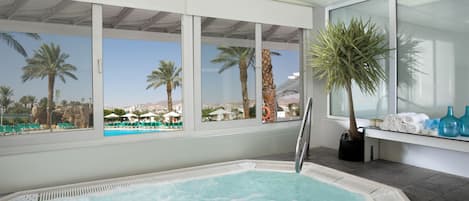 A spa area with a hot tub, large windows, and a view of a pool and palm trees.