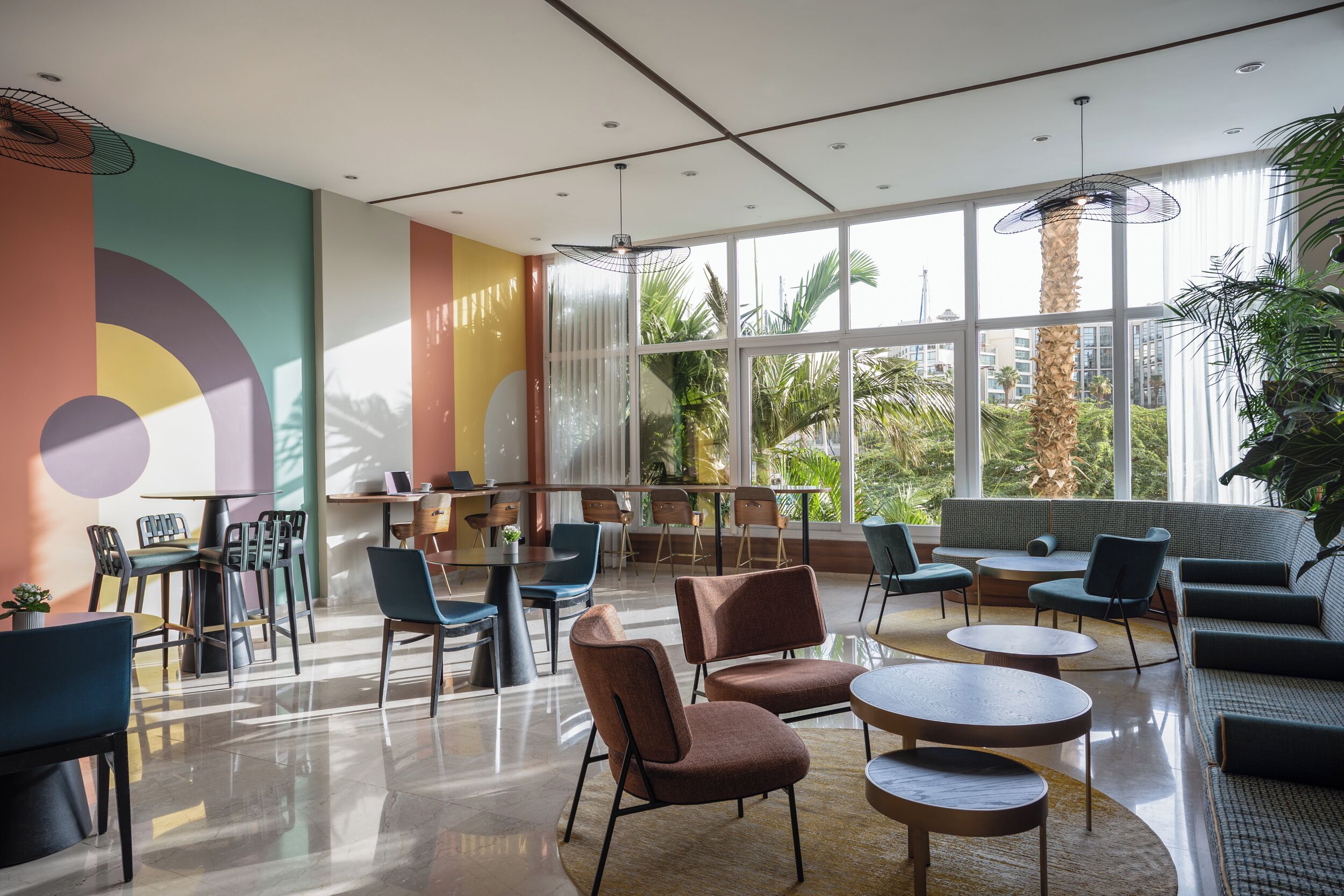 A modern lounge area with a mix of seating options, large windows offering views of palm trees, and a colorful abstract wall design.