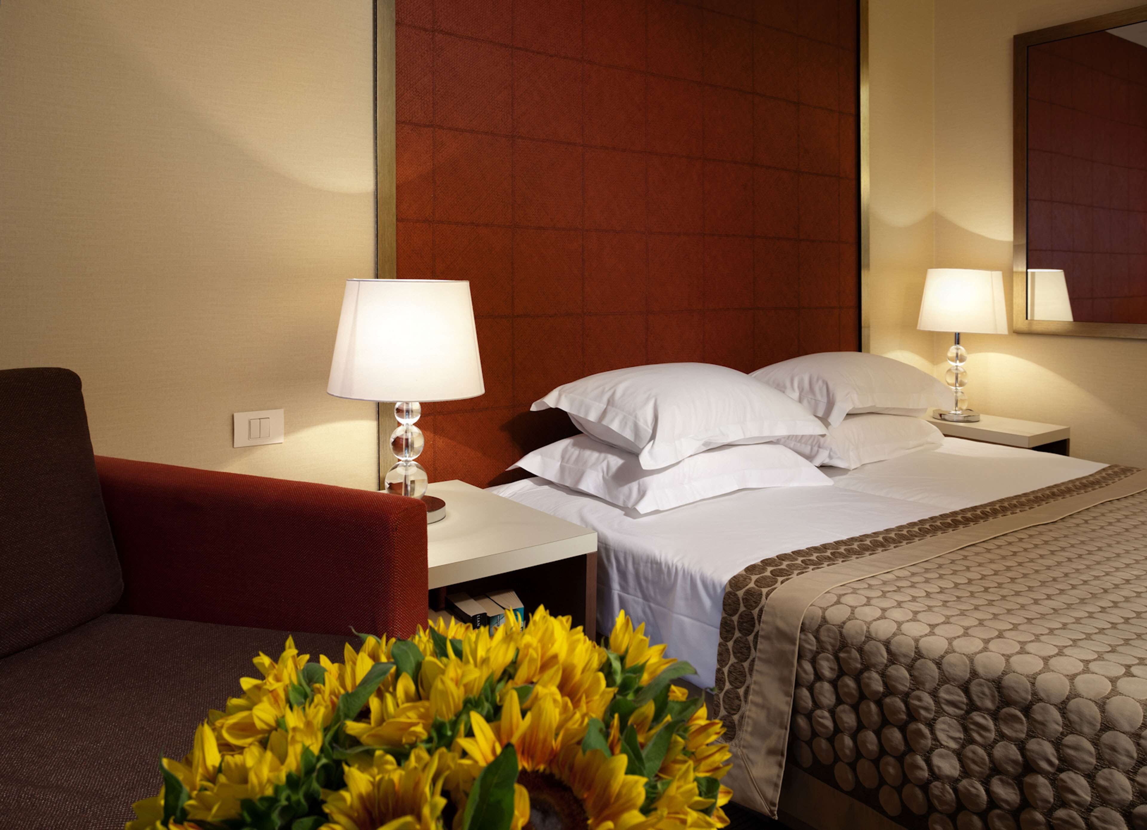 A hotel room with a bed, two bedside tables, a red armchair, and a vase of yellow flowers.
