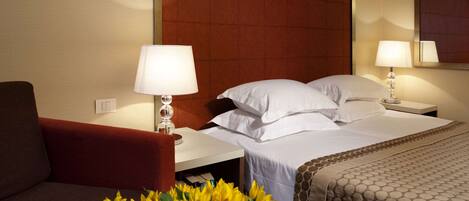 A hotel room with a bed, two bedside tables, a red armchair, and a vase of yellow flowers.
