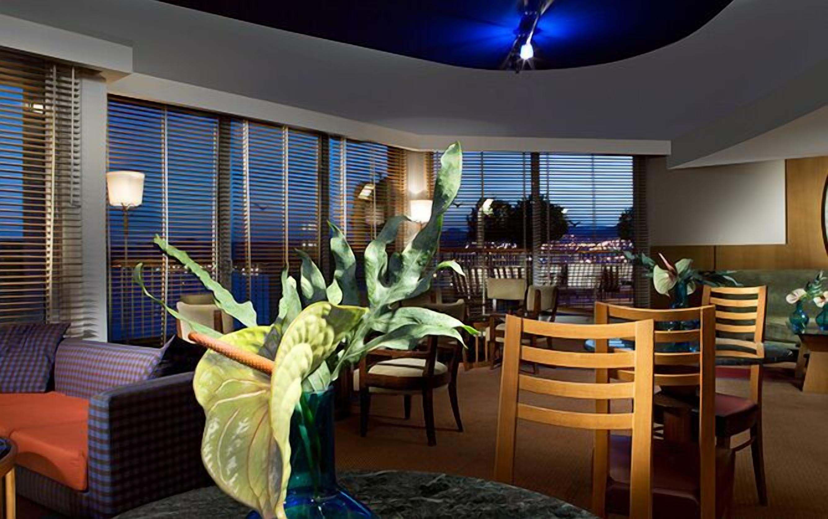 Una sala de estar moderna con comedor, ventanas amplias con persianas y vista a un paisaje urbano al atardecer.