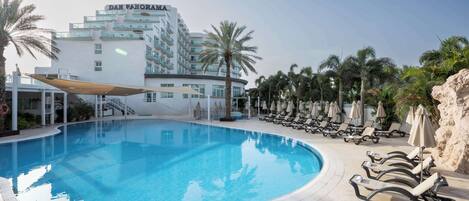 Una piscina con sillas de descanso y sombrillas, rodeada de palmeras y un hotel llamado "Dan Panorama".