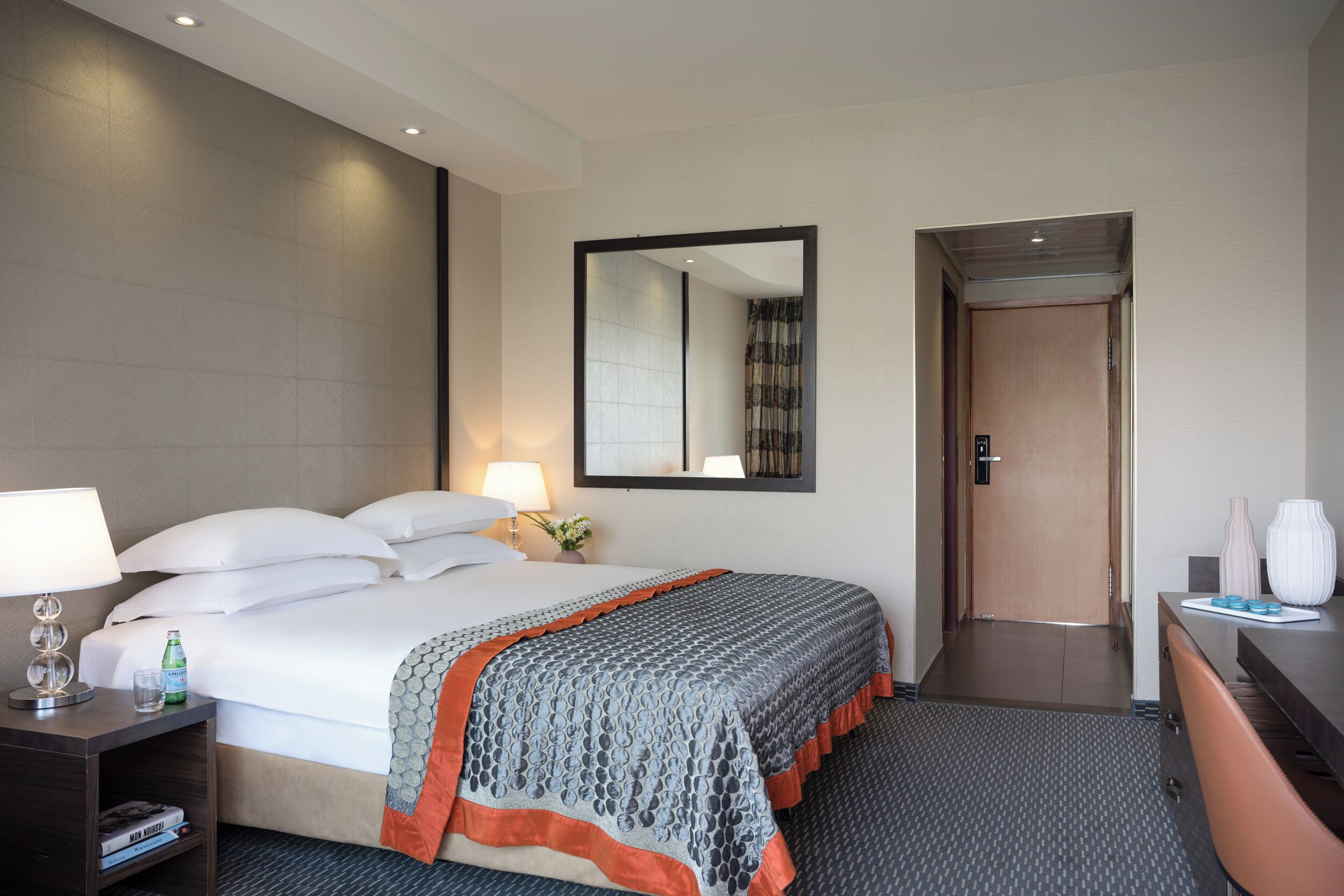 Una habitación de hotel con una cama bien hecha, una mesita de noche con una lámpara y una botella, un aparador con un jarrón y un espejo en la pared.
