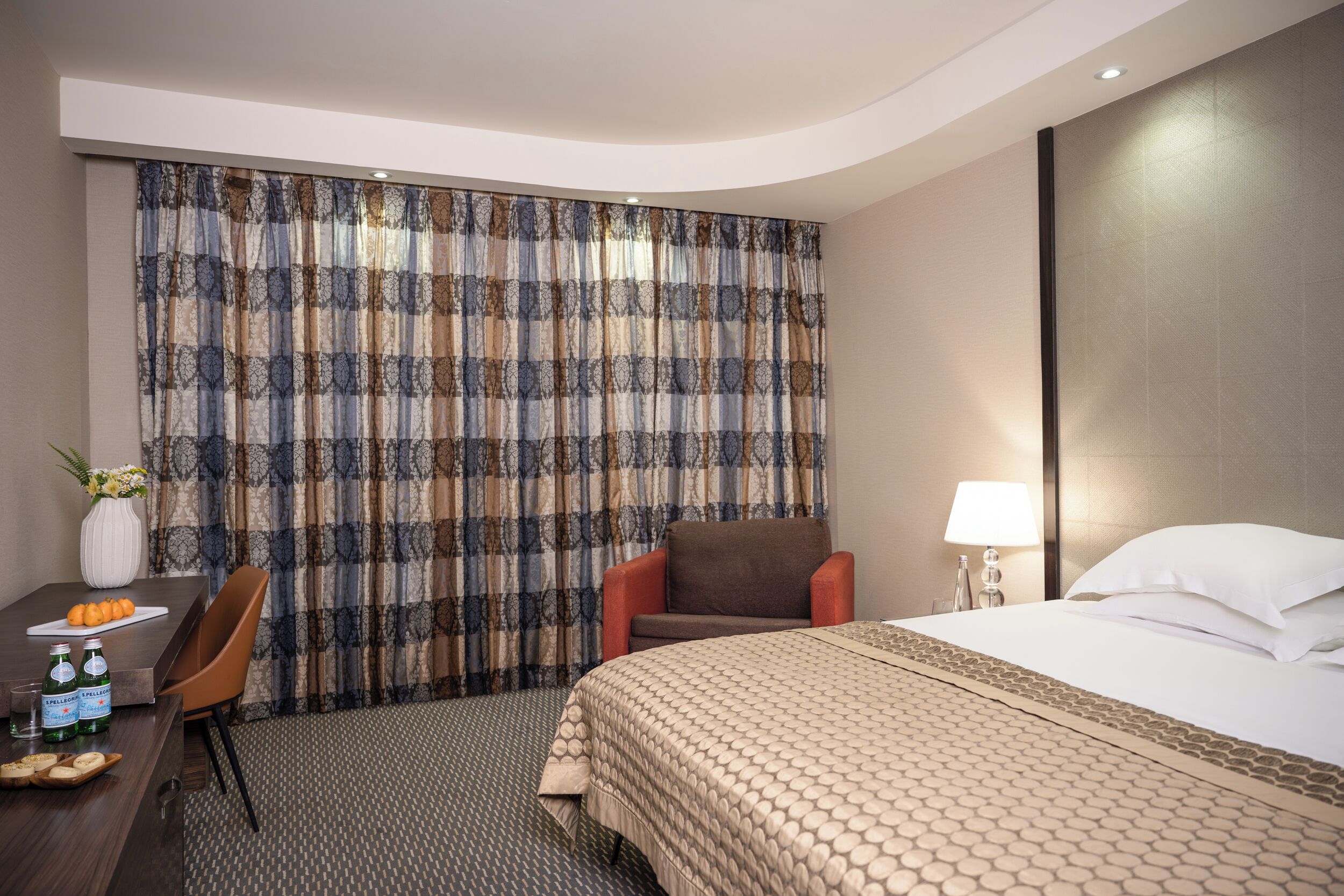 Habitación de hotel con una cama, una silla, un escritorio con un florero y botellas, y una cortina estampada.