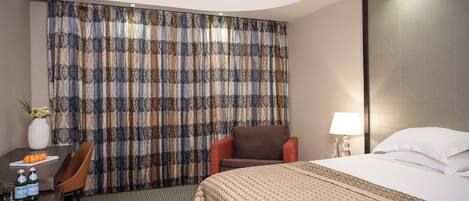 Habitación de hotel con una cama, una silla, un escritorio con un florero y botellas, y una cortina estampada.