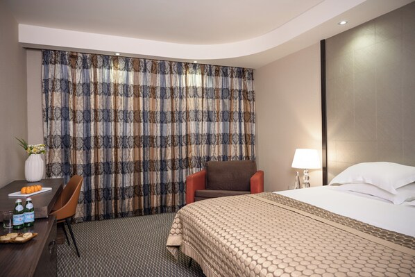 A hotel room with a bed, a chair, a desk with a vase and bottles, and a patterned curtain.