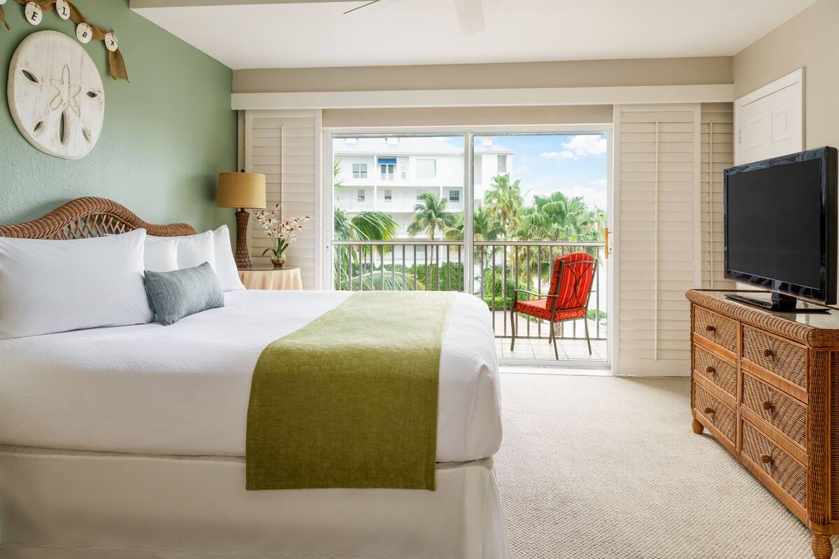 Une chambre d’hôtel équipée d’un lit, d’une télévision, d’une commode et d’un balcon offrant une vue sur les palmiers.