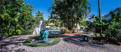 A paved walkway with a decorative fountain, surrounded by lush greenery and palm trees.