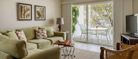 Un salón con un sofá, una silla de mimbre, una mesa con una lámpara, un televisor y un ventanal corredizo que da a un patio exterior con mesa y sillas.
