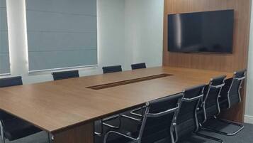 A conference room with a long wooden table, black chairs, a flat-screen TV mounted on the wall, and a large window with blinds.
