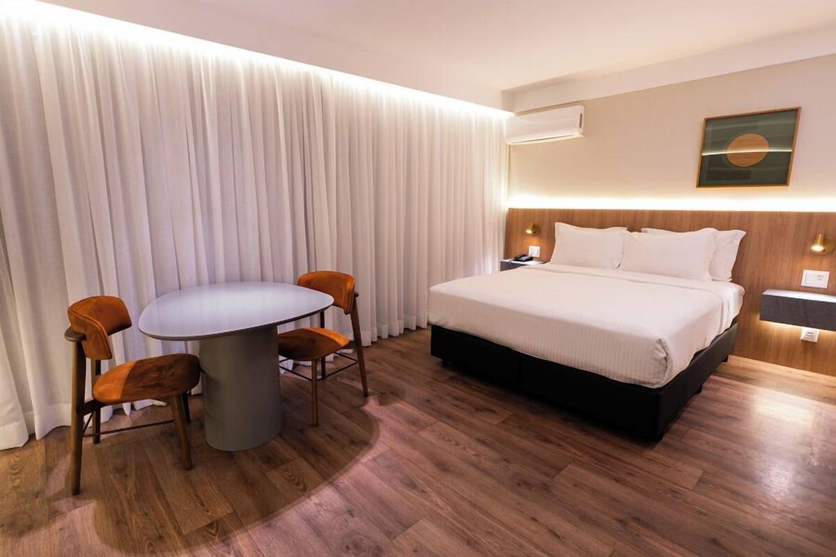 A hotel room with a bed, a round table with chairs, a large window with curtains, and a wall-mounted air conditioner.