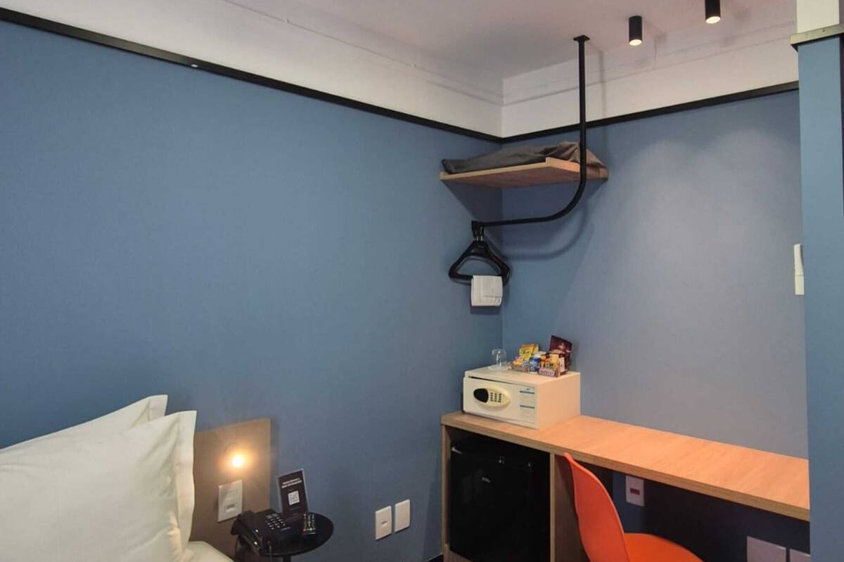 A compact hotel room with a bed, a desk, a chair, and a small kitchenette.
