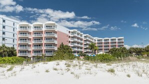 Un hotel frente a la playa con varios balcones, una playa arenosa al frente y un cielo azul despejado.