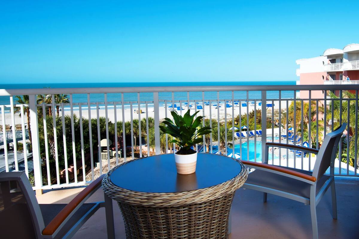 A balcony with a wicker table and chair, a potted plant, and a view of the beach and pool area.