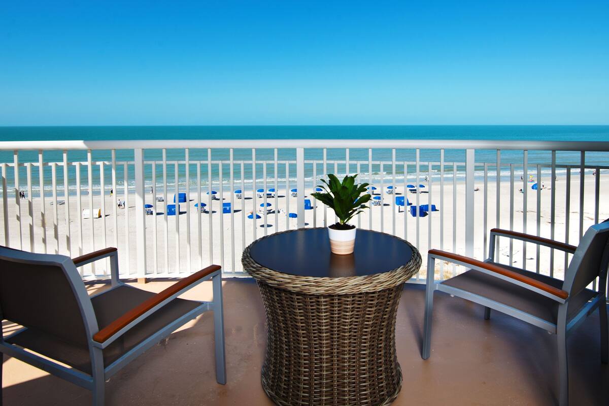A balcony with wicker chairs, a table, and a potted plant, overlooking a beach with blue umbrellas.