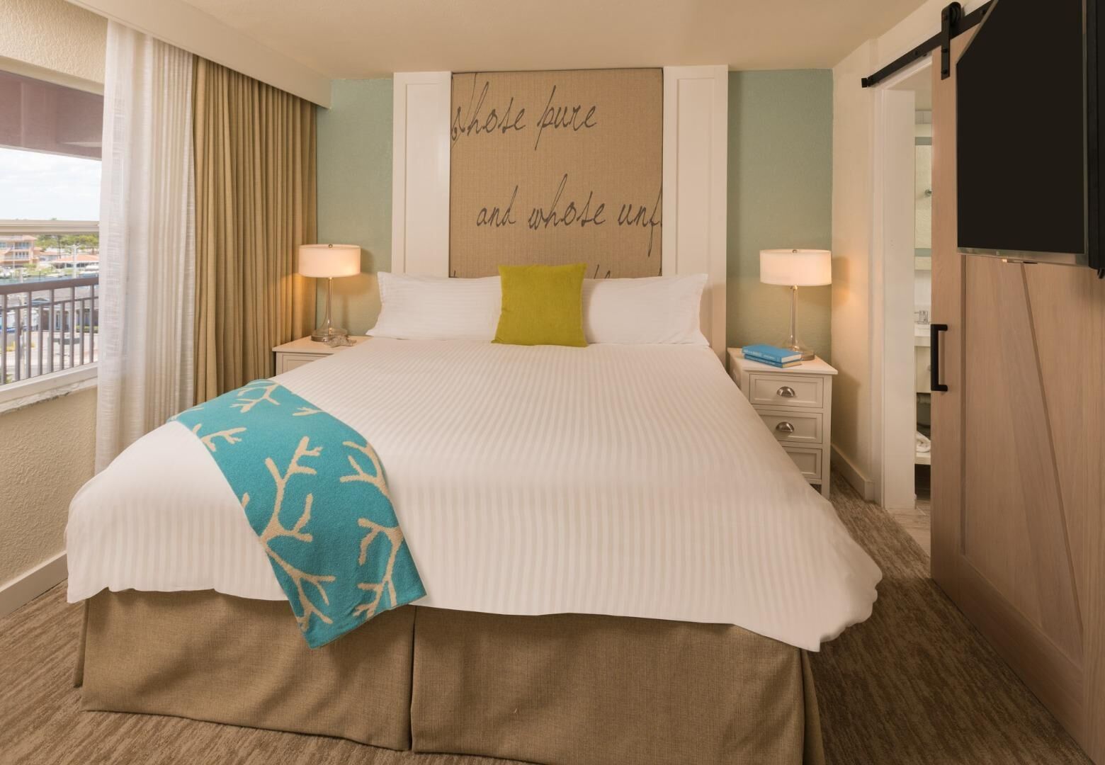 A hotel room with a large bed, two bedside tables with lamps, a view of the outside, and a wall with a quote.