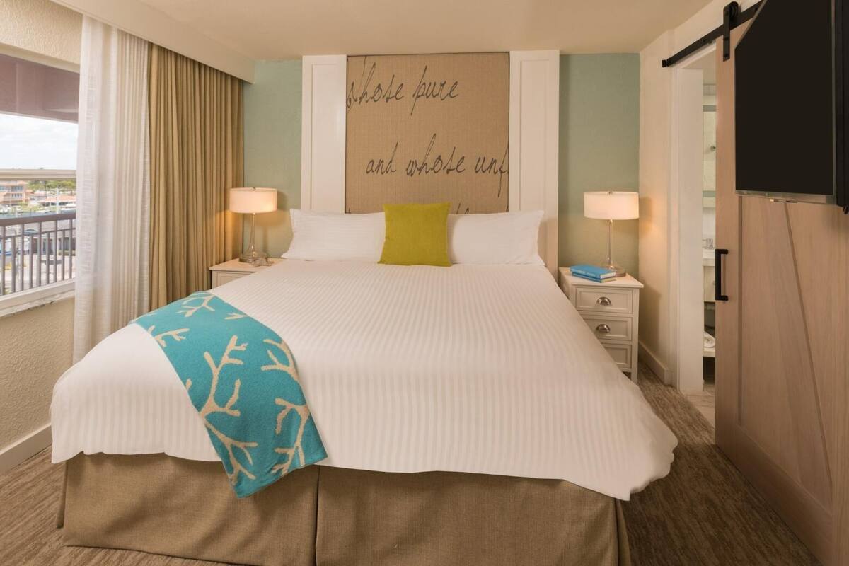A hotel room with a large bed, two bedside tables with lamps, a view of the outside, and a wall with a quote.