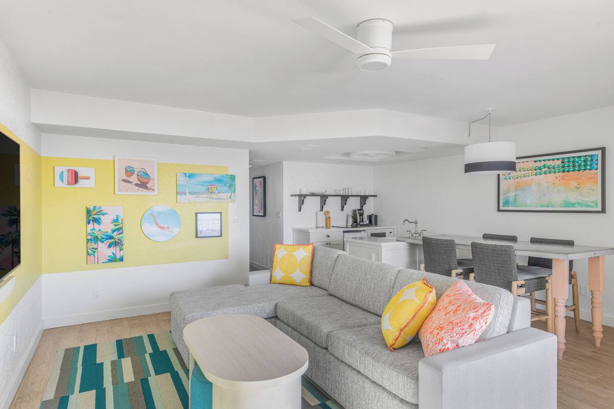 A modern living room with a gray sofa, yellow walls, and artwork. There is a dining area with a table and chairs.