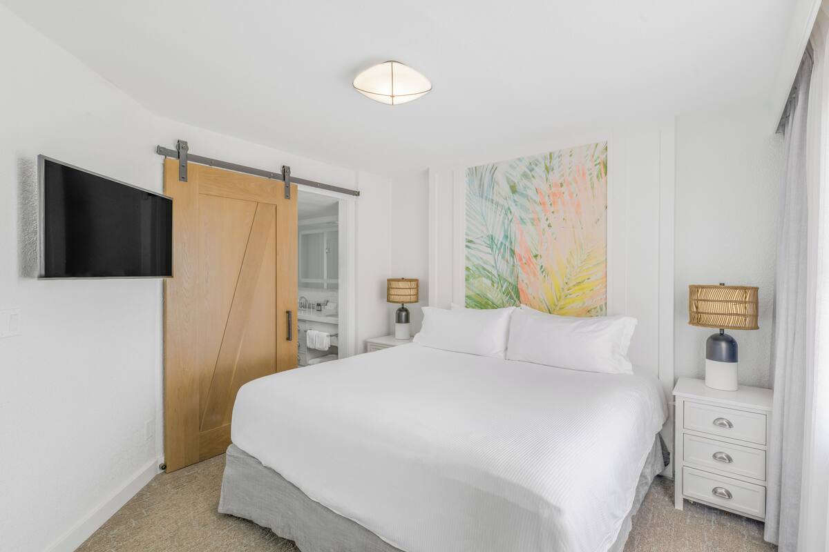 A bedroom with a bed, a TV mounted on the wall, a wooden door, a bedside table with a lamp, and a colorful artwork on the wall.