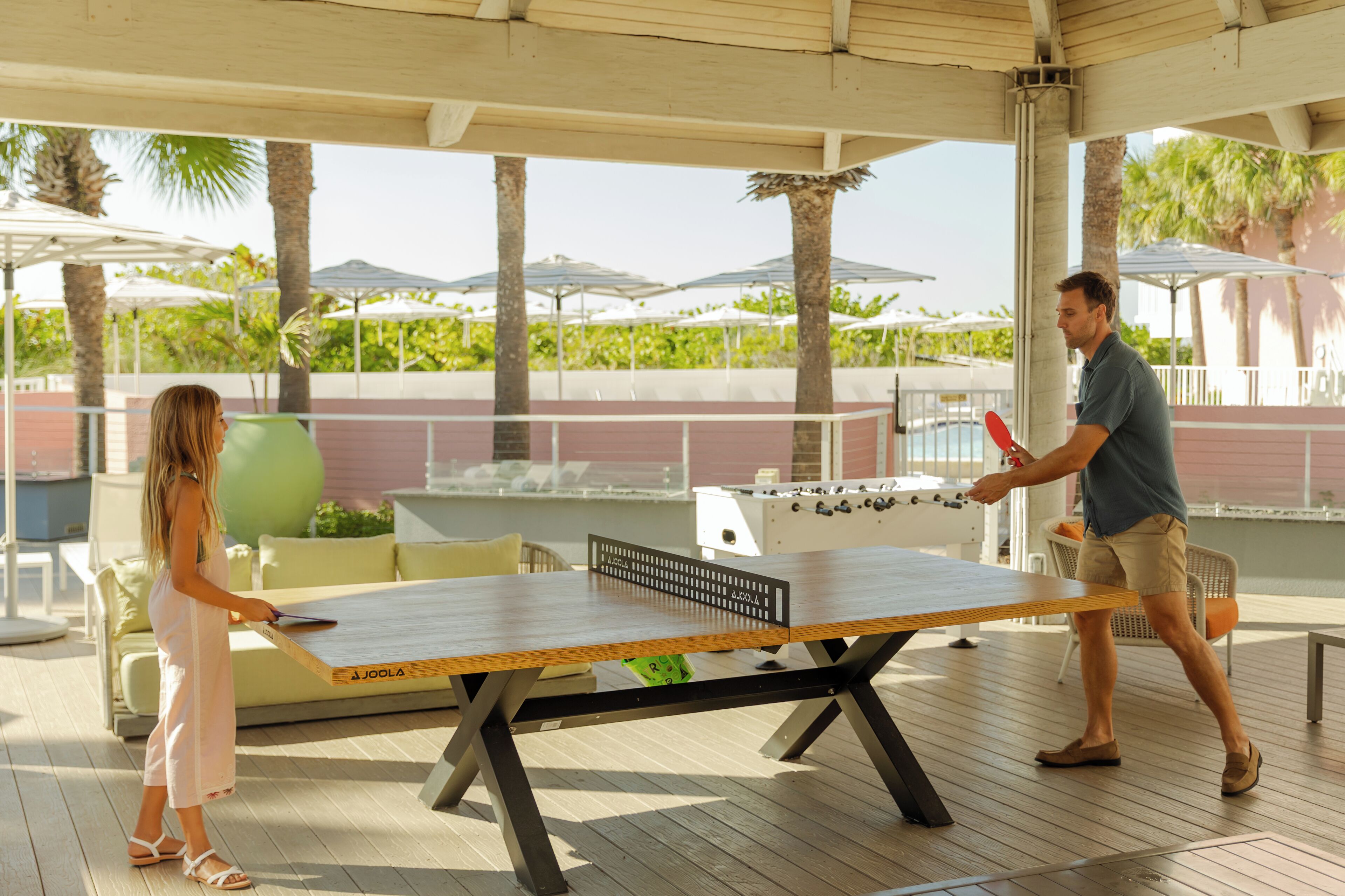A table tennis table with a net in a covered outdoor area with palm trees and umbrellas in the background.