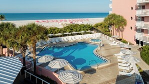 Zona de piscina con sillones y sombrillas, una playa al fondo y un edificio con balcones.