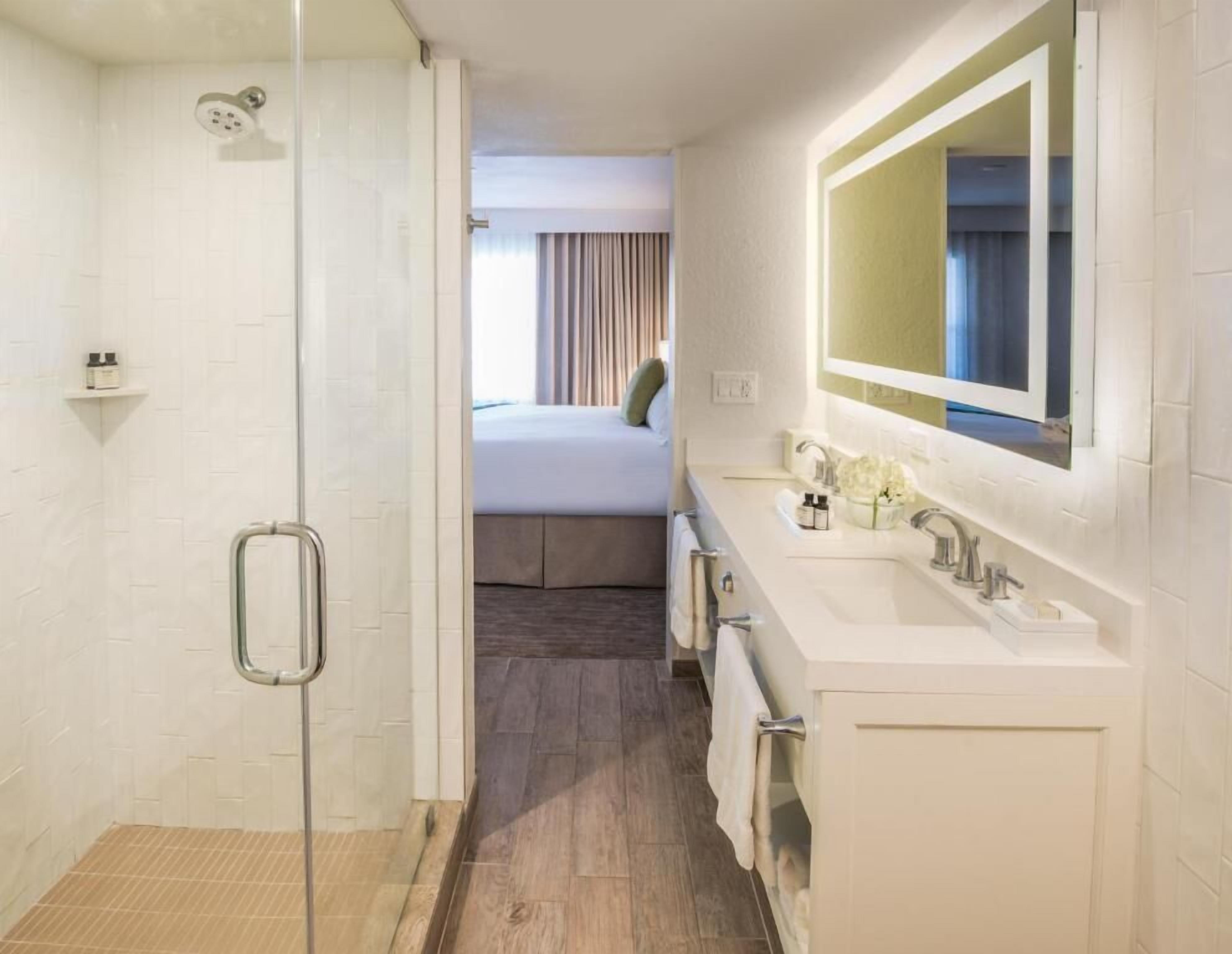 A hotel bathroom with a shower, a bed, a sink, and a mirror.