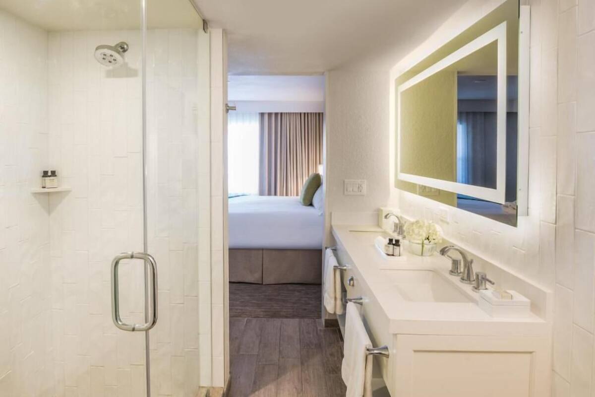 A hotel bathroom with a shower, a bed, a sink, and a mirror.