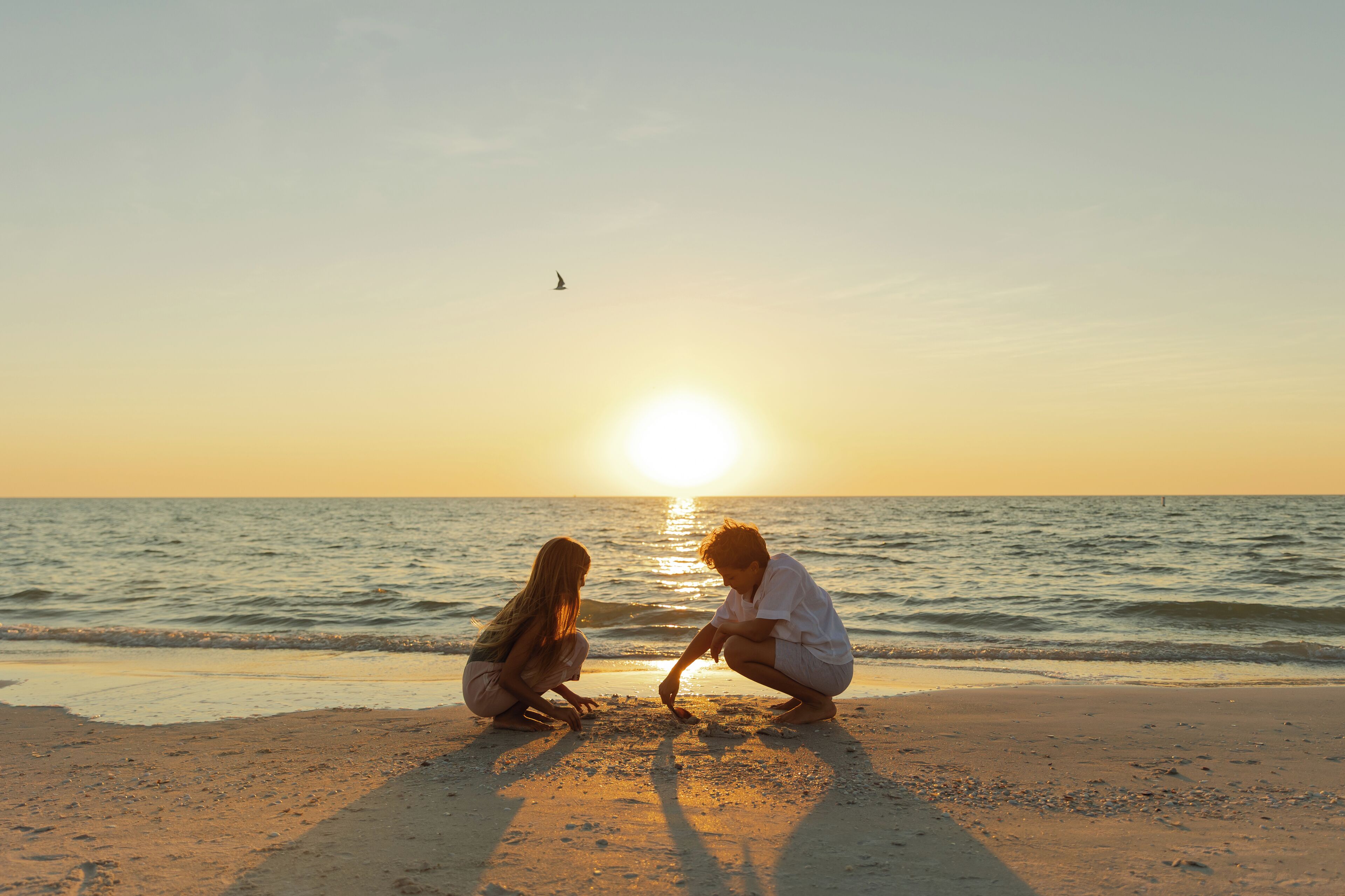Escena de playa con dos personas sentadas sobre la arena mirando al océano durante el atardecer.