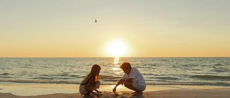 Escena de playa con dos personas sentadas sobre la arena mirando al océano durante el atardecer.