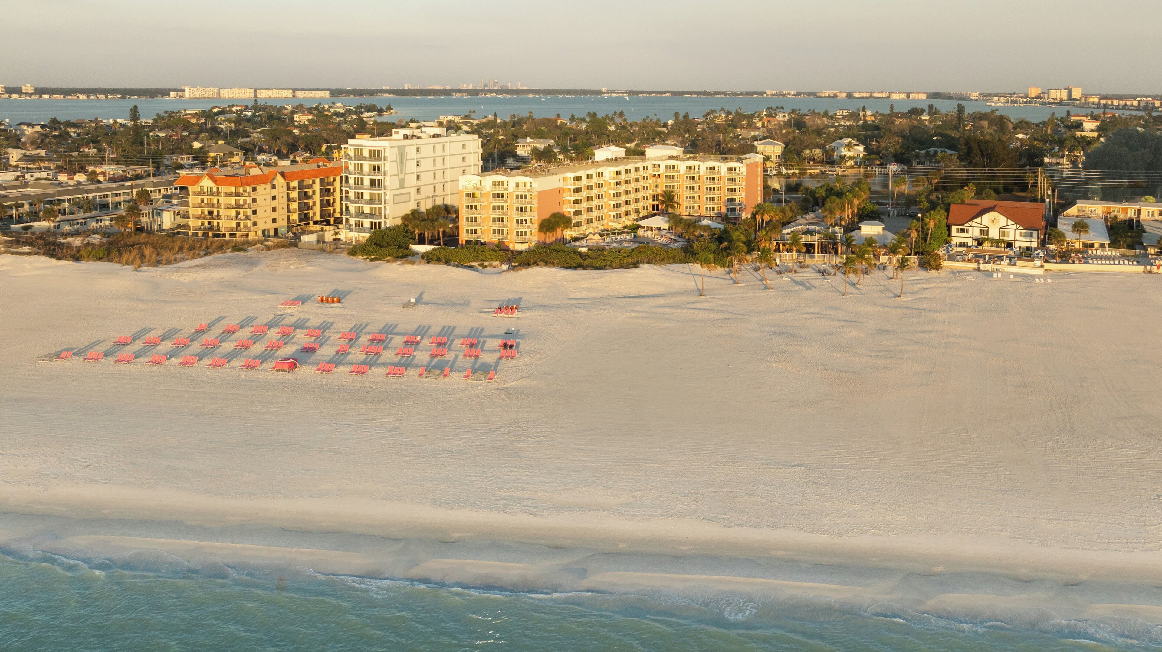A beachfront hotel with multiple buildings, a sandy beach with rows of umbrellas, and a view of the ocean and distant city skyline.